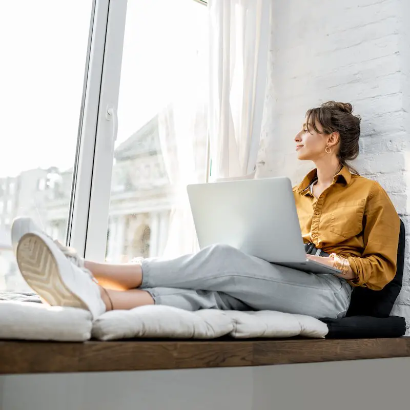 Woman Working On Her Laptop Sitting By The Window Sill, Digital Nomad Concept